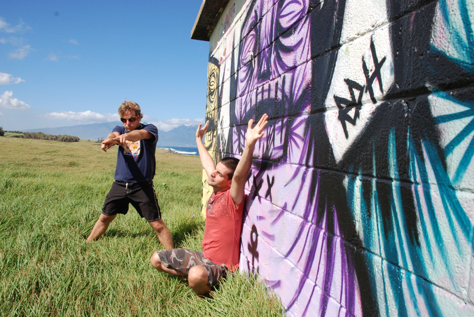 Dust of Vraj: two band members against a graffiti-painted wall in a green Maui field, ocean and mountains behind