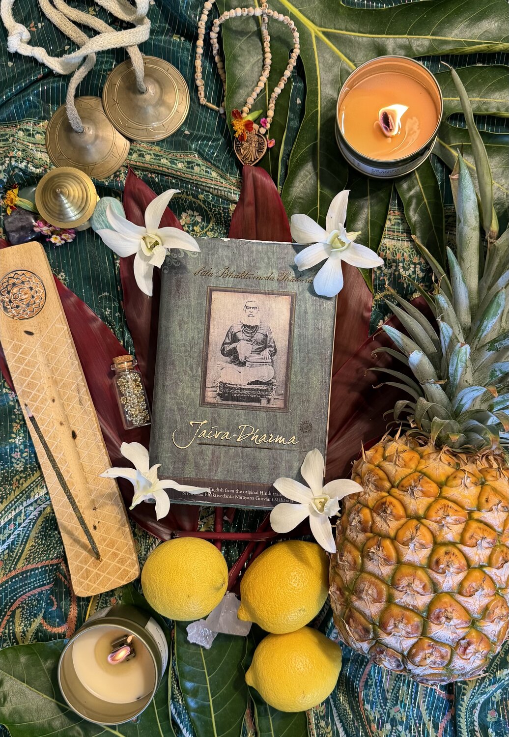 The Jaiva Dharma book resting on ti leaves among orchids, lit candles, karatalas, japa beads, incense, pineapple and lemons — an altar offering